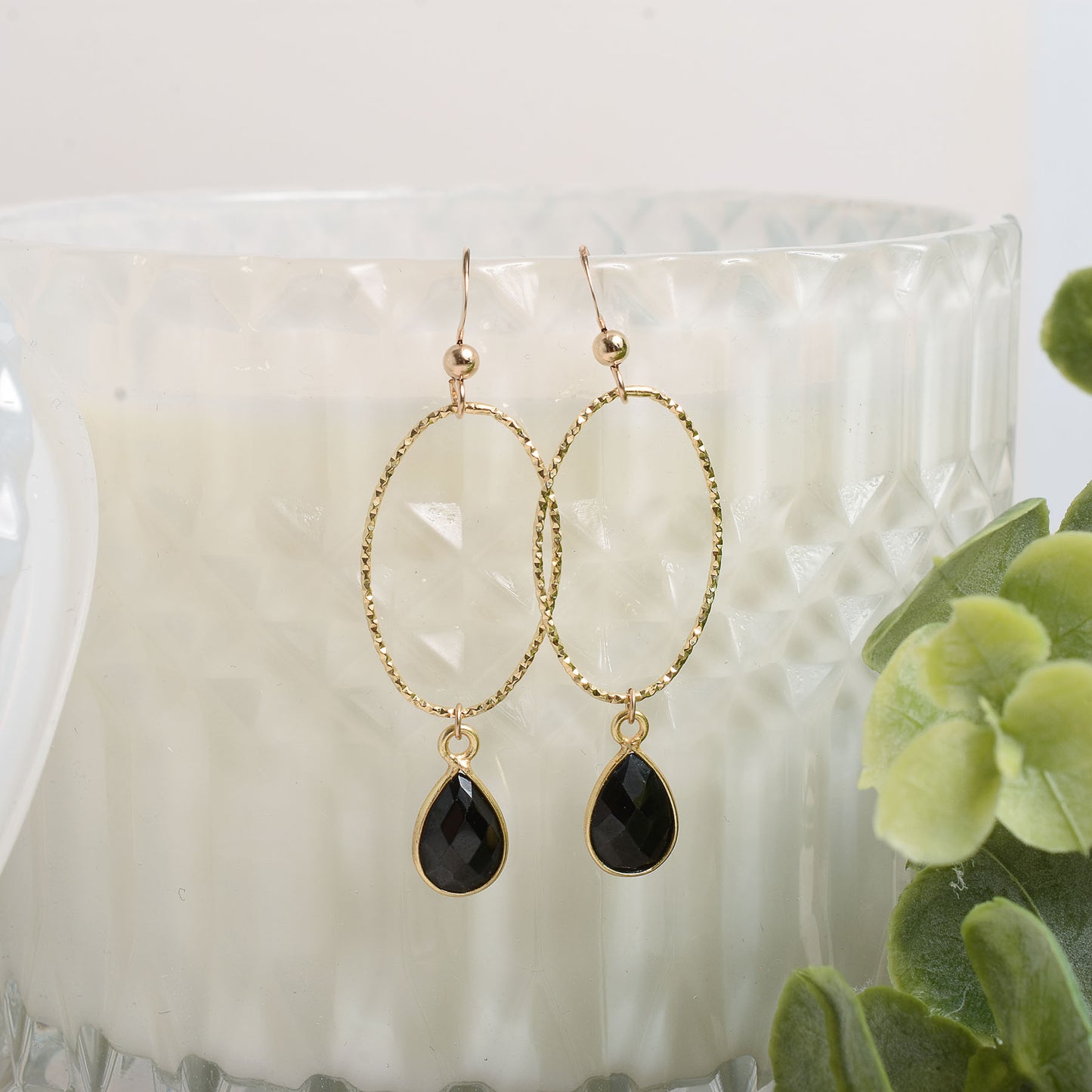 Gold earrings with black teardrop charms on a white surface with green leaves.
