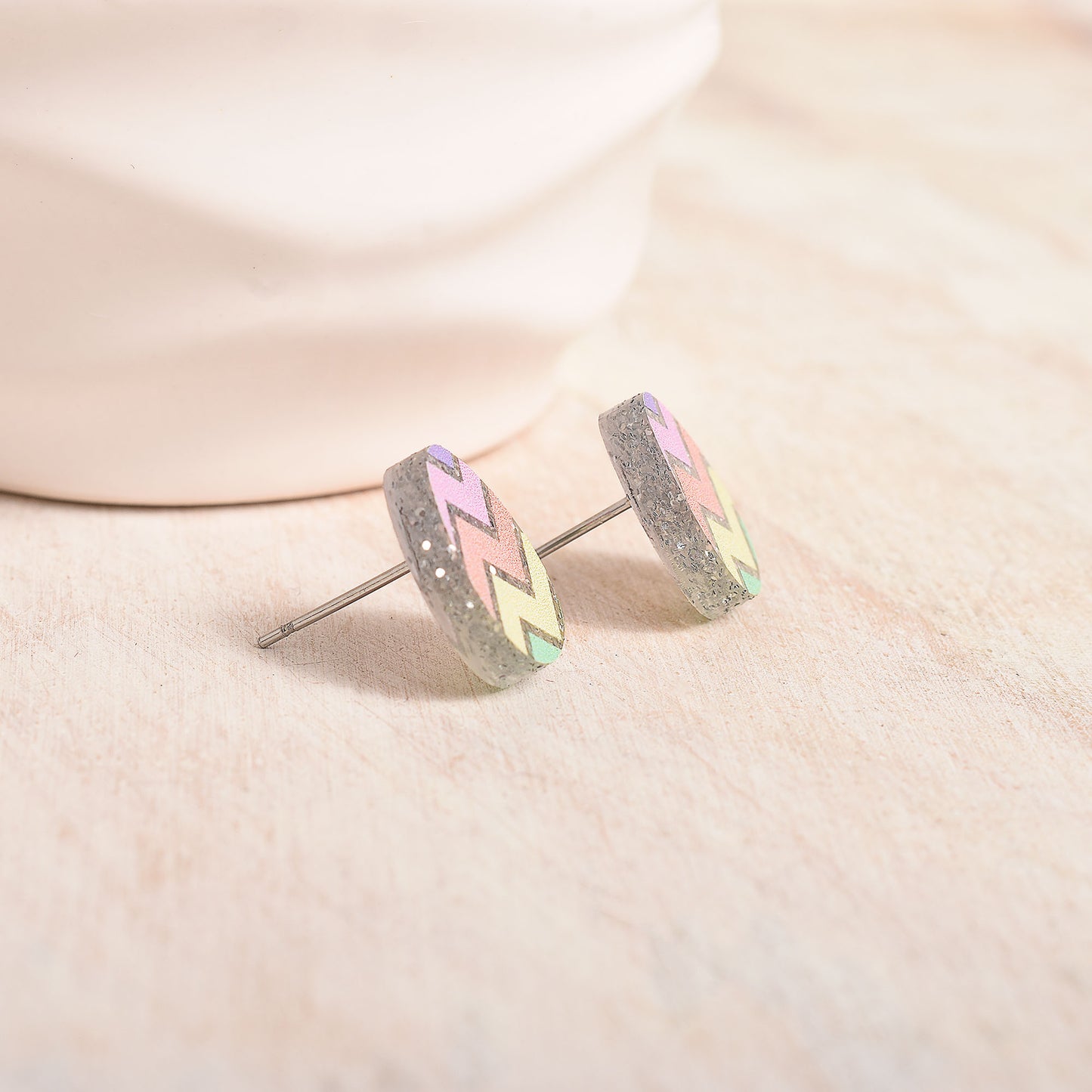 A pair of silver stud earrings with a glittery gray background, decorated with a chevron pattern in pastel pink, green, and yellow, resting on a light beige textured surface.