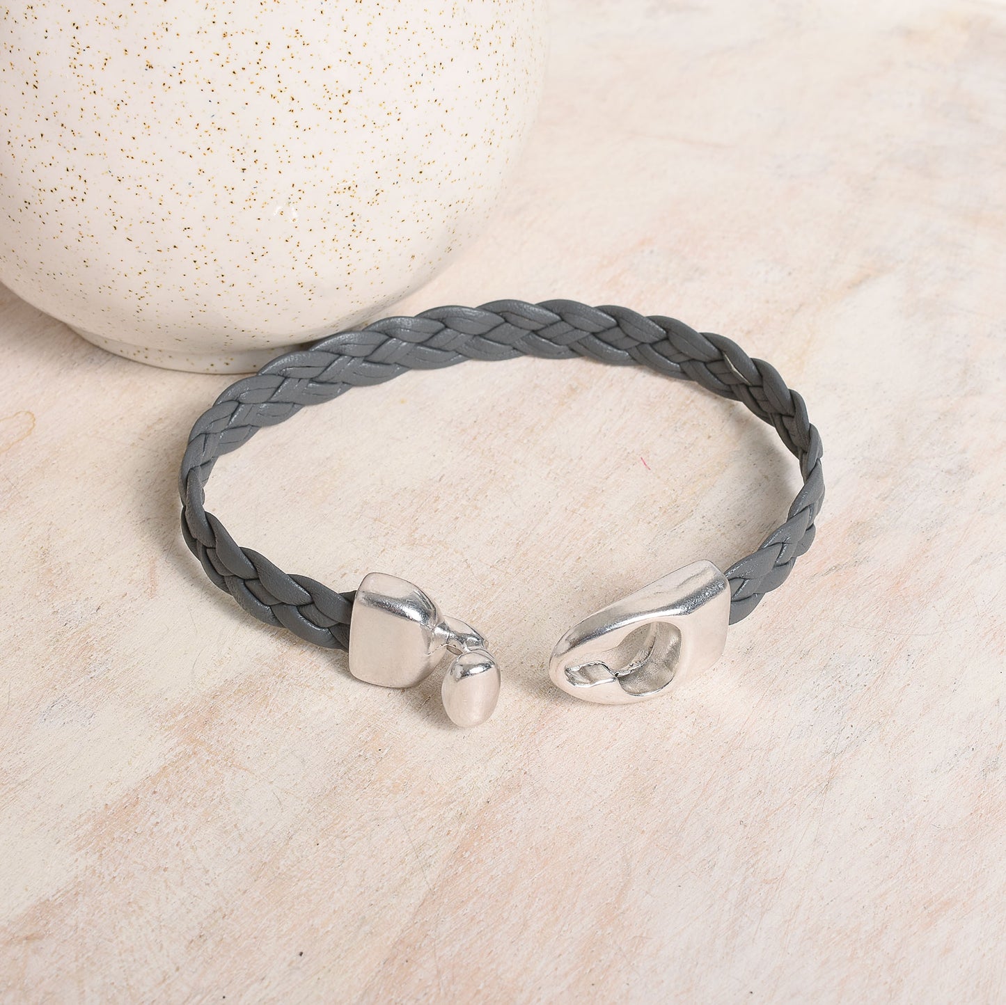 A gray braided leather bracelet with polished silver-tone toggle clasp, displayed on a light beige surface.