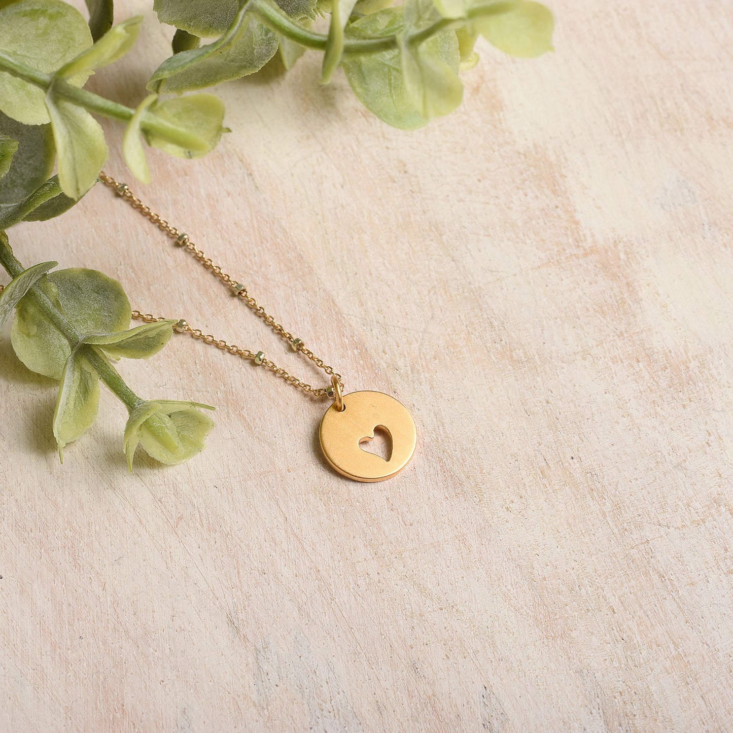 Gold necklace with a round pendant on a beige surface with green leaves.