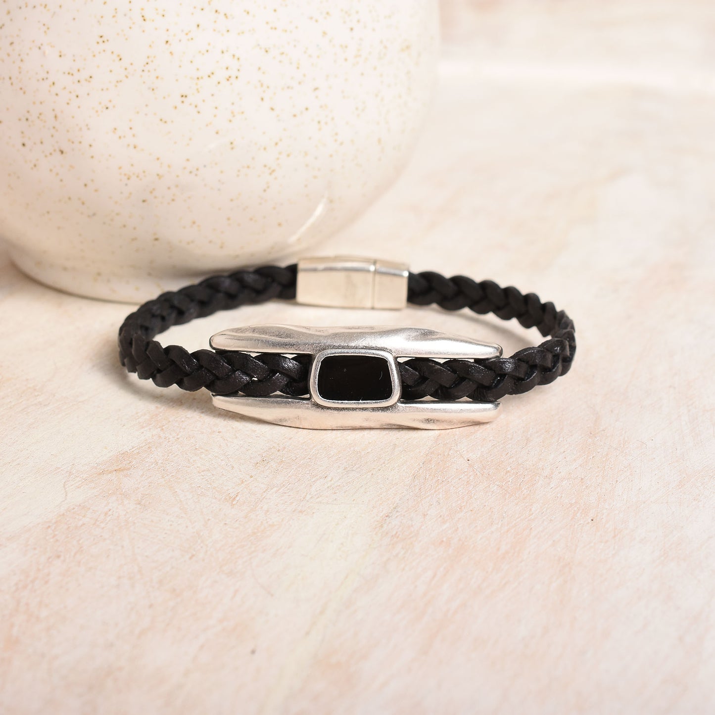 A black braided leather bracelet with a polished silver-toned statement bar and a small black epoxy inlay displayed on a light background.