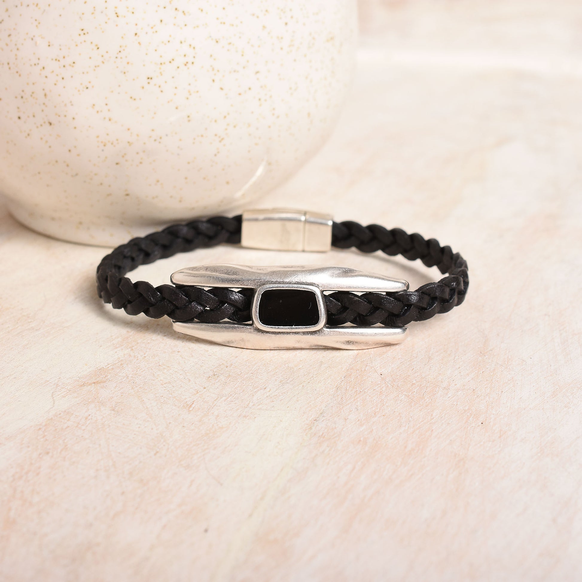 A black braided leather bracelet with a polished silver-toned statement bar and a small black epoxy inlay displayed on a light background.