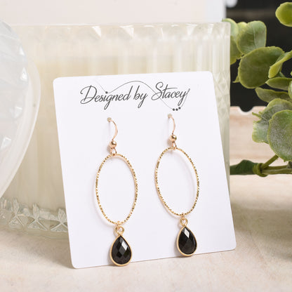 Gold earrings with black teardrop drops on a 'Designed by Stacey' card with a candle and plant in the background.