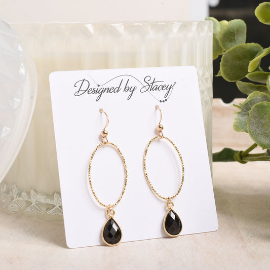 Gold earrings with black teardrop drops on a 'Designed by Stacey' card with a candle and plant in the background.