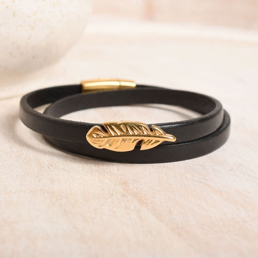 Black leather bracelet with a gold feather charm on a beige background