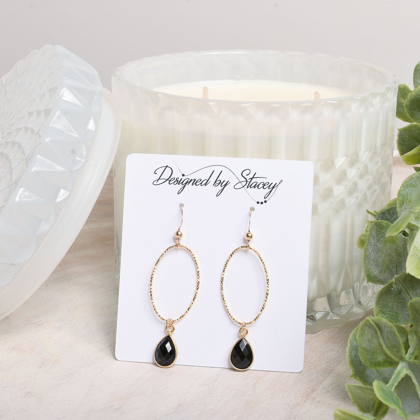 Gold earrings with black stones on a card labeled 'Designed by Stacey' in front of a candle.