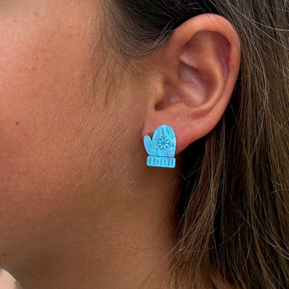 A close-up of a person's ear wearing a light blue stud earring shaped like a mitten with a snowflake design.