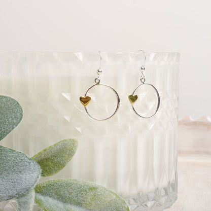Bronze heart and silver circle earrings on an opaque stand with a plant in the background.