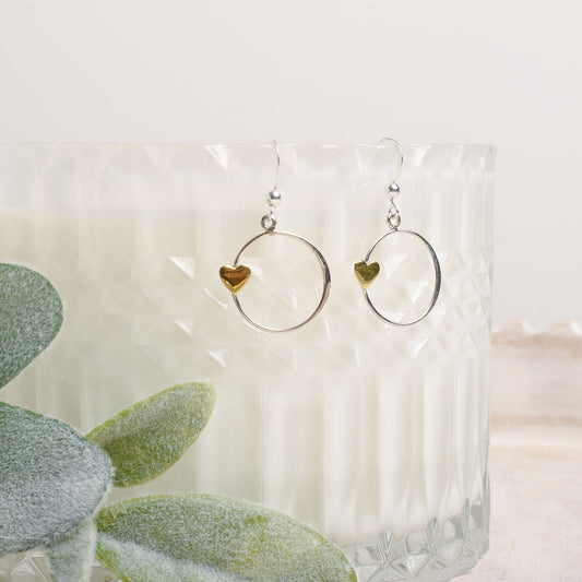 Bronze heart and silver circle earrings on an opaque stand with a plant in the background.