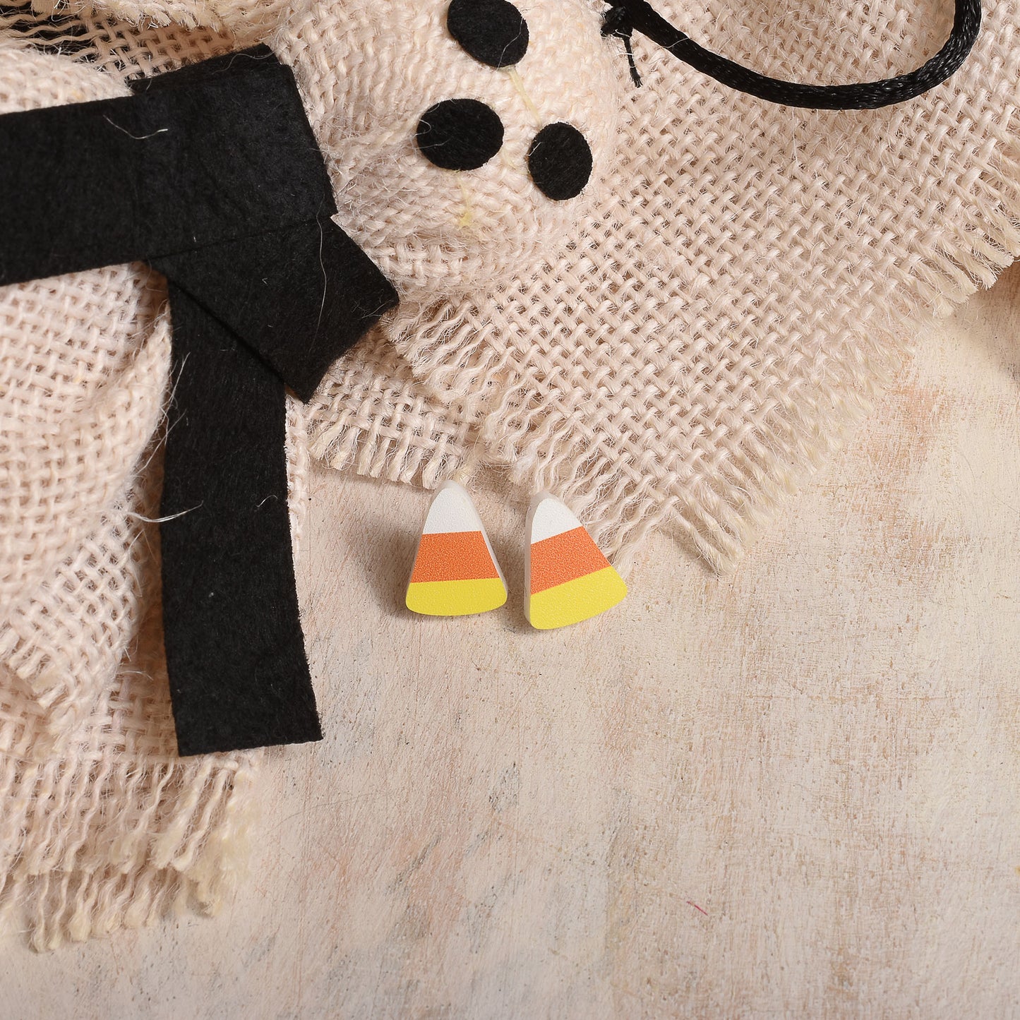 Candy corn stud earrings with white, orange, and yellow sections, displayed on a rustic burlap surface next to a black felt bow and Halloween-themed props.