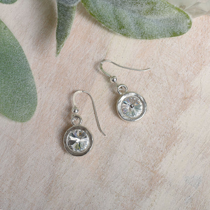 Silver earrings with clear Austrian crystals on a textured surface with green leaves.