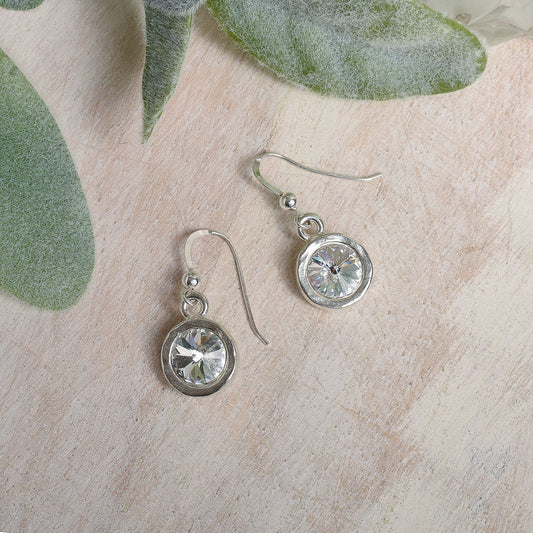 Silver earrings with clear Austrian crystals on a textured surface with green leaves.