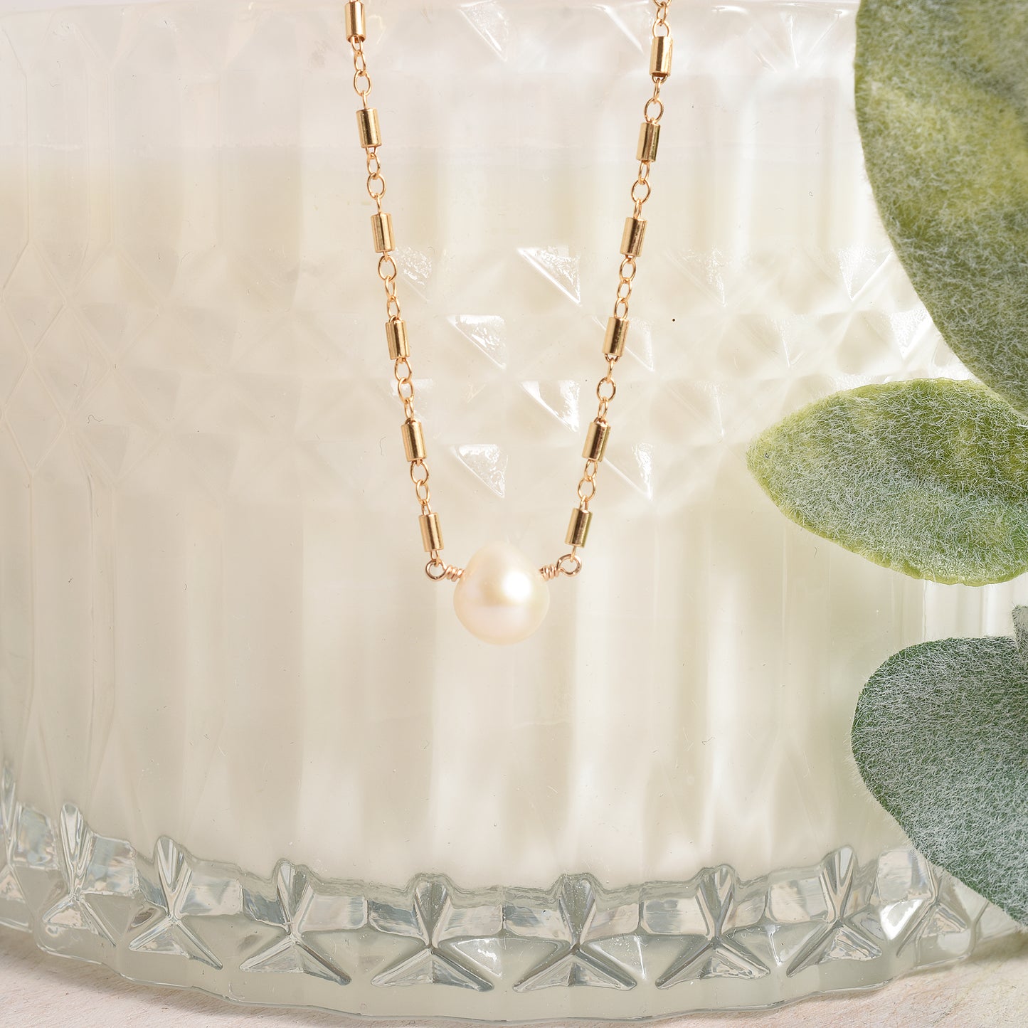 Gold necklace with a pearl pendant on a textured surface with green leaves.