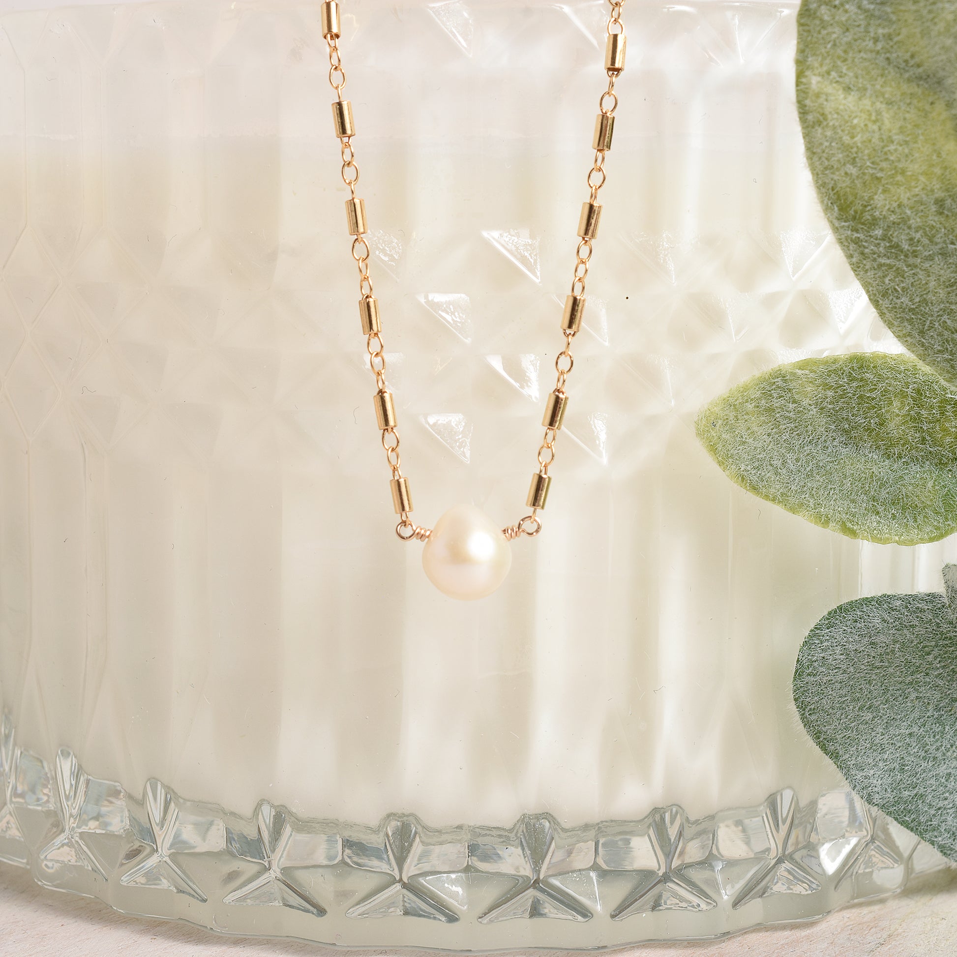 Gold necklace with a pearl pendant on a textured surface with green leaves.