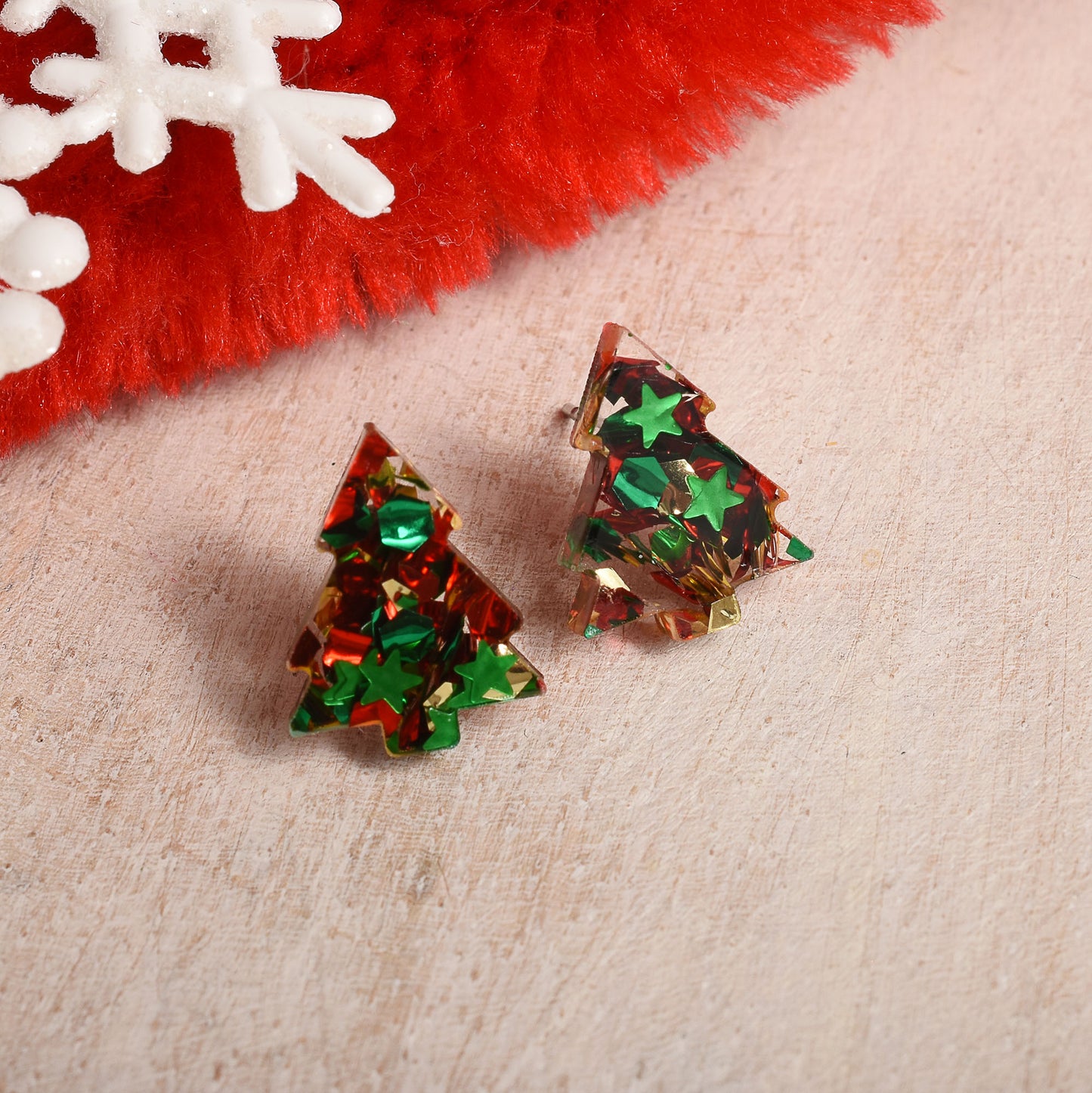 Two Christmas tree-shaped stud earrings filled with green and red glitter, displayed on a textured beige surface with a festive red and white fuzzy prop in the background.