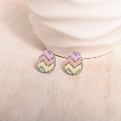 A pair of pastel chevron Easter egg earrings with glittery silver edges, resting on a textured beige surface.