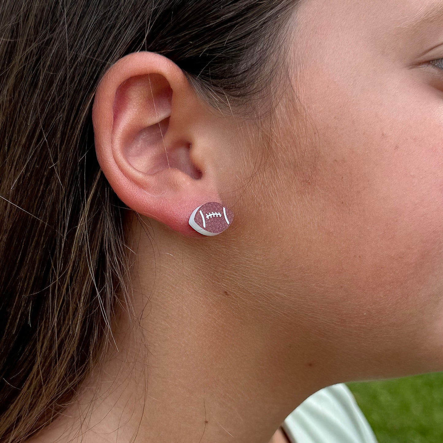 A close-up side profile of a model's ear wearing a small stud earring shaped like a football in brown and white.