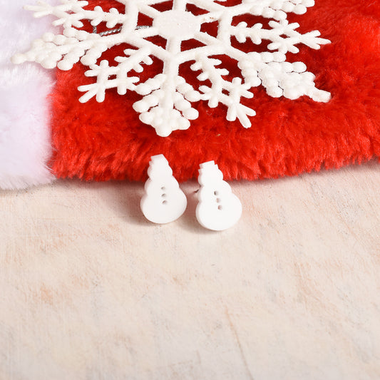 A pair of small white snowman-shaped earrings displayed on a red fuzzy surface with a white snowflake and a cream-colored textured background.