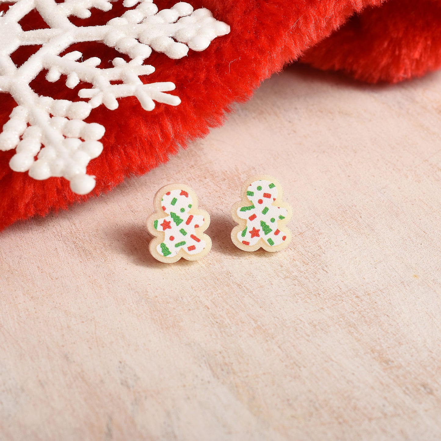 A pair of small gingerbread man stud earrings decorated with green, red, and white sprinkles, resting on a light beige textured surface next to a red fuzzy fabric and a white snowflake ornament.