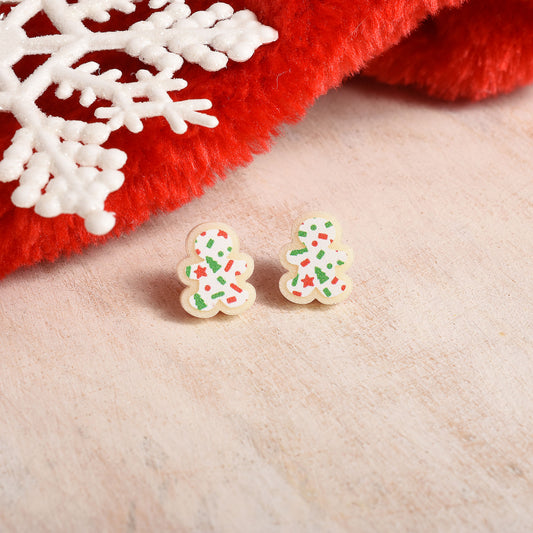 A pair of small gingerbread man stud earrings decorated with green, red, and white sprinkles, resting on a light beige textured surface next to a red fuzzy fabric and a white snowflake ornament.
