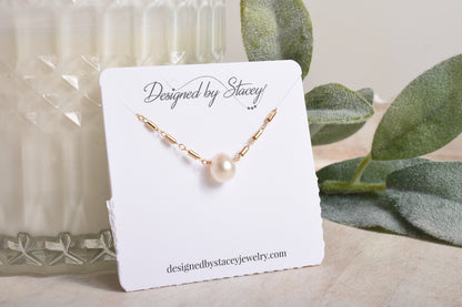 Gold necklace with a pearl pendant on a branded card, with green leaves in the background.
