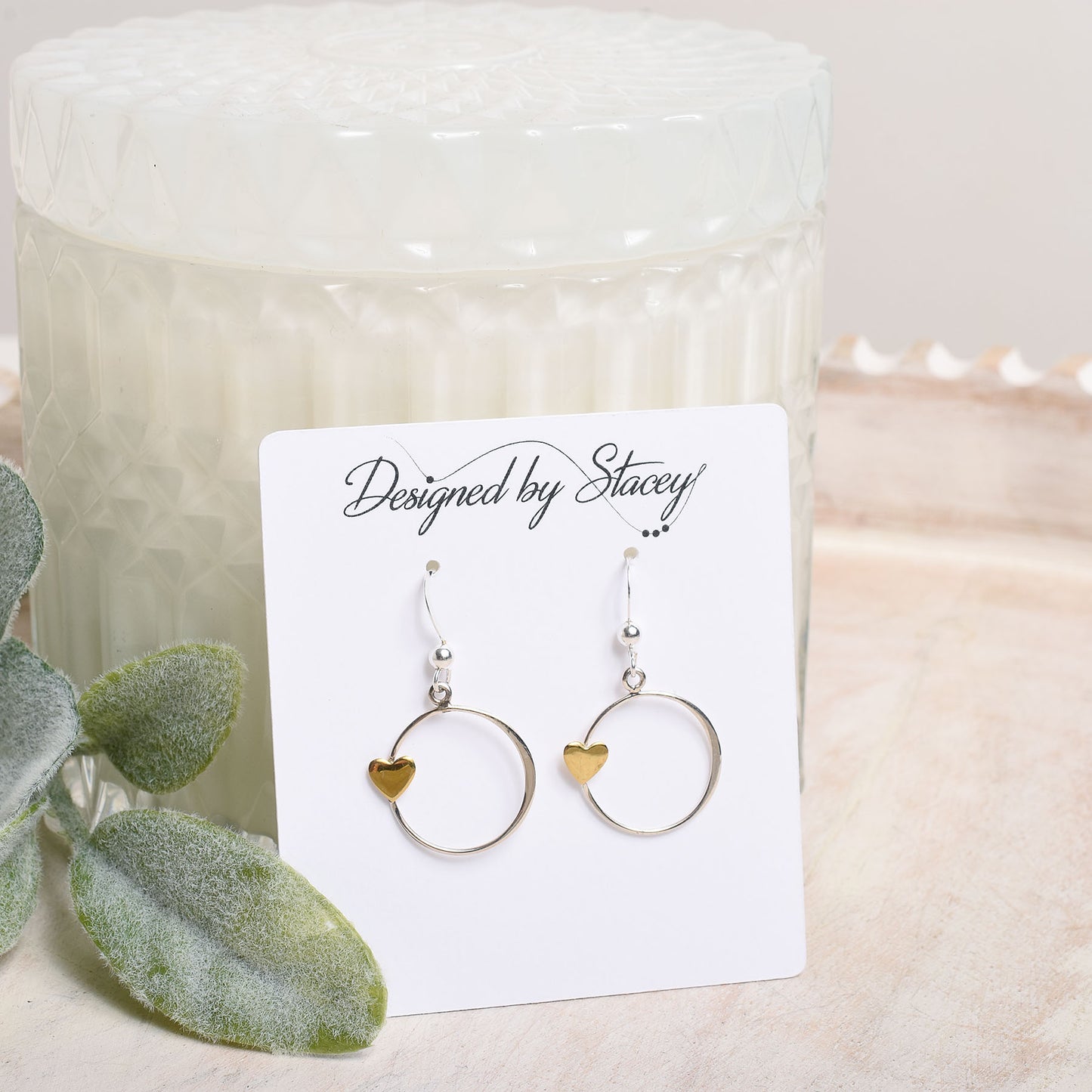 Pair of earrings on a card labeled 'Designed by Stacey' with a candle and plant in the background.