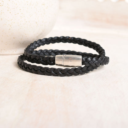 A black braided leather wrap bracelet with a silver-toned metal clasp, resting on a light beige textured surface.