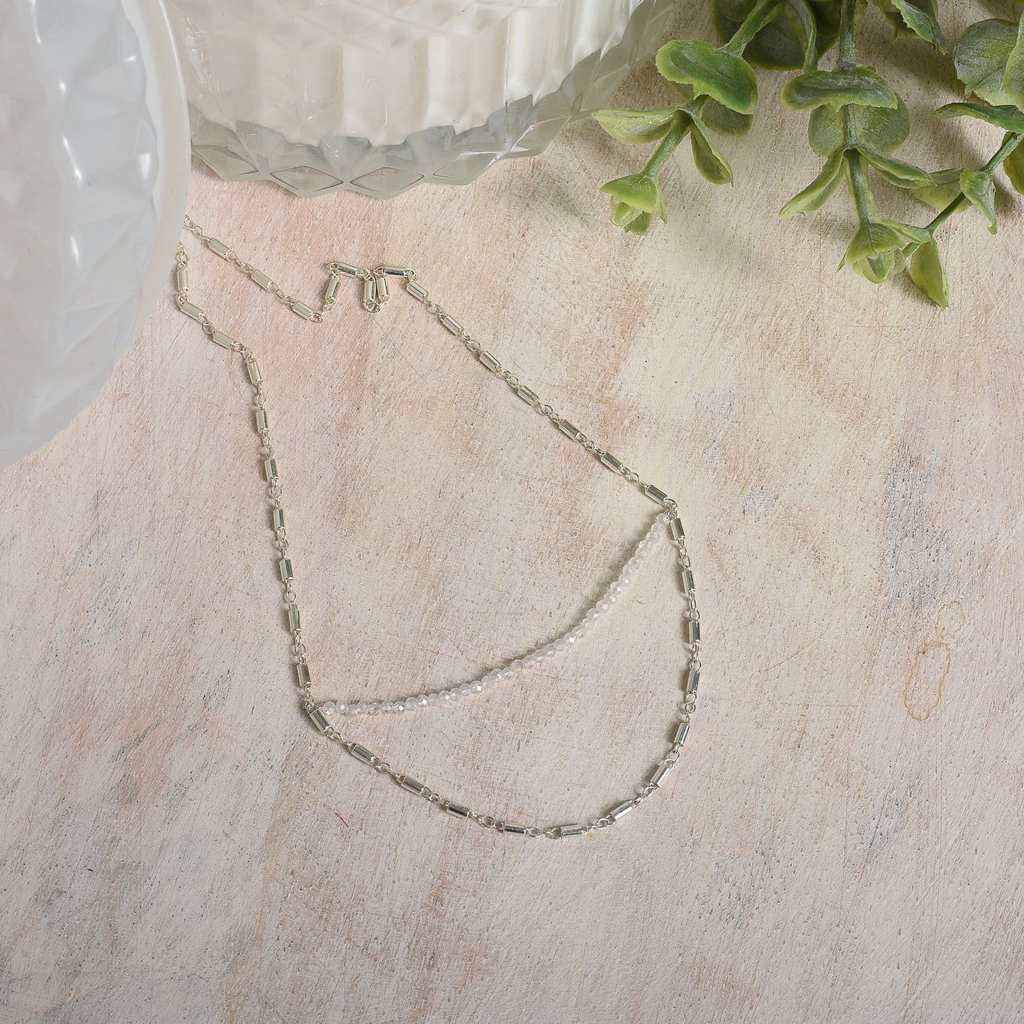 Silver necklace on a light wooden surface with green leaves in the corner