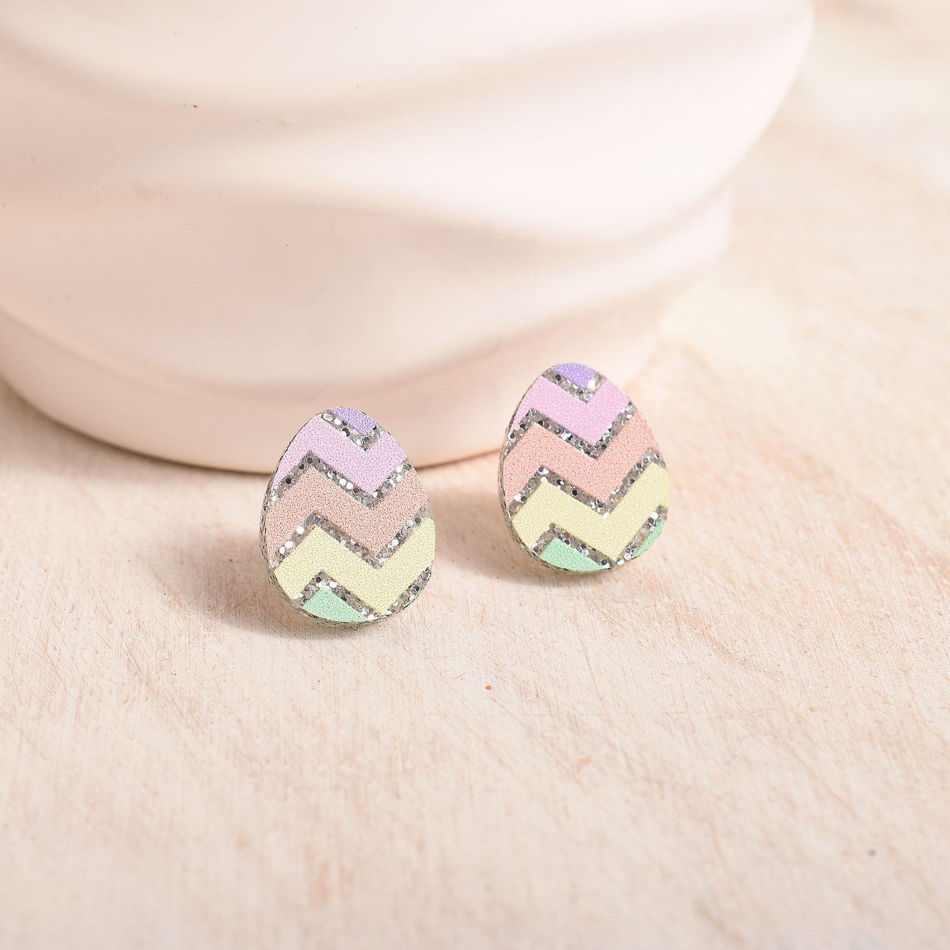 A pair of pastel chevron Easter egg stud earrings with glittery silver edges, resting on a light beige textured surface in front of a cream ceramic object.