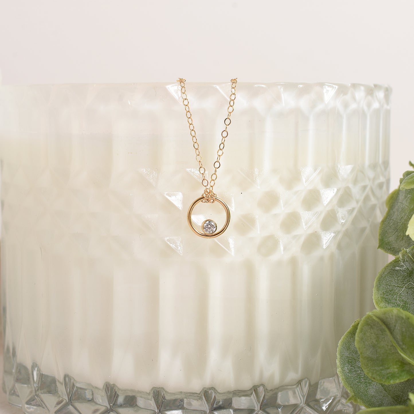 Gold necklace with a small round pendant on a textured white surface with a plant in the corner.