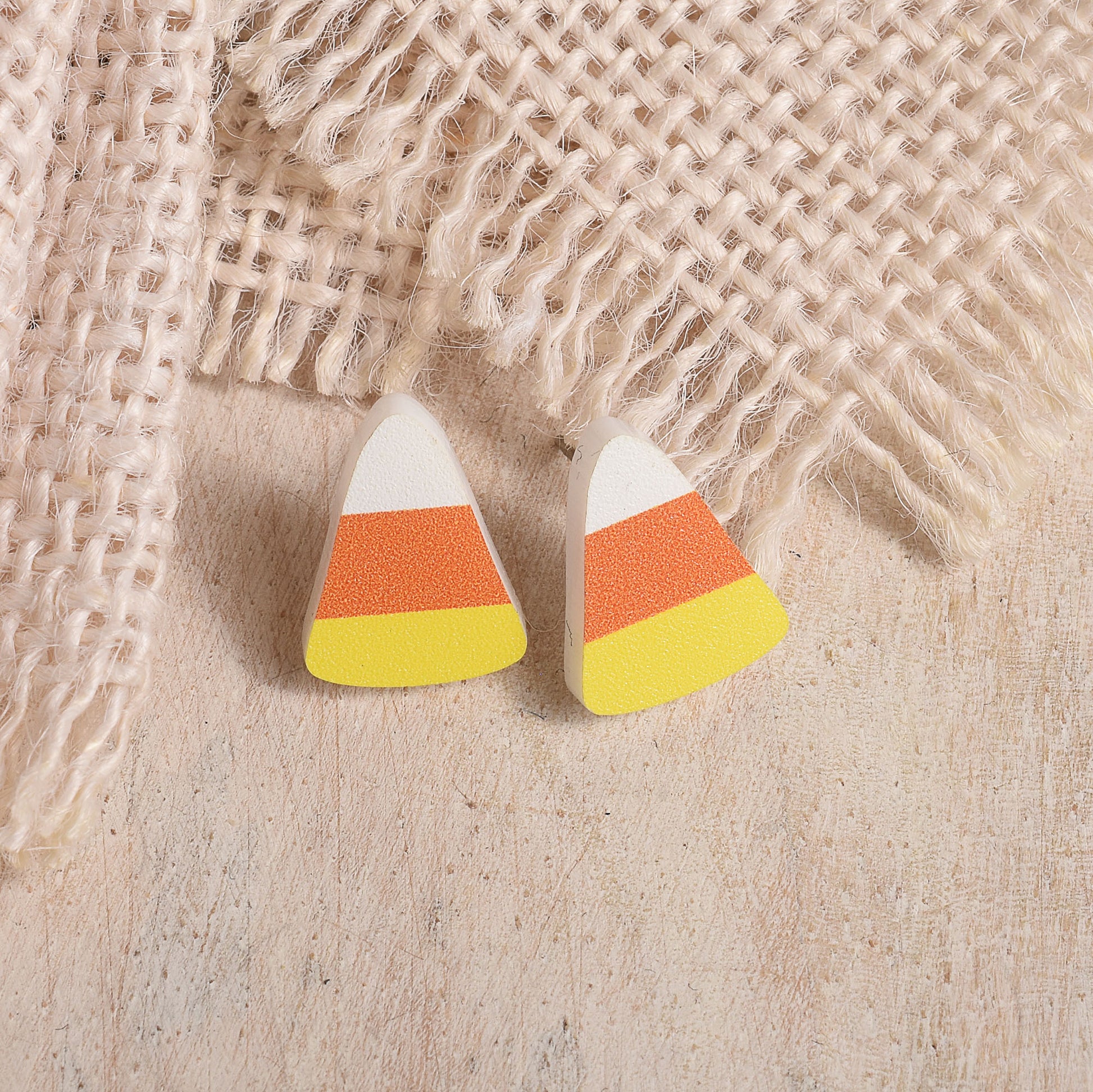 Candy corn shaped stud earrings with white, orange, and yellow stripes, displayed on a light wood surface with a burlap fabric backdrop.