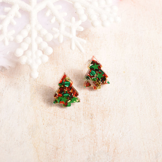 Two small Christmas tree-shaped stud earrings filled with glittery red and green resin, placed on a light cream surface with a white decorative snowflake prop in the background.