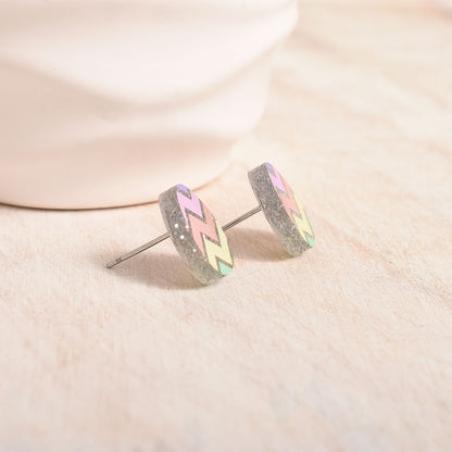 A pair of silver stud earrings with a glittery gray background, decorated with a chevron pattern in pastel pink, green, and yellow, resting on a light beige textured surface.