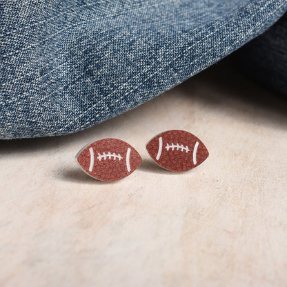 A pair of football-shaped stud earrings with a brown finish and white laces, displayed on a light beige surface next to a folded blue denim fabric.