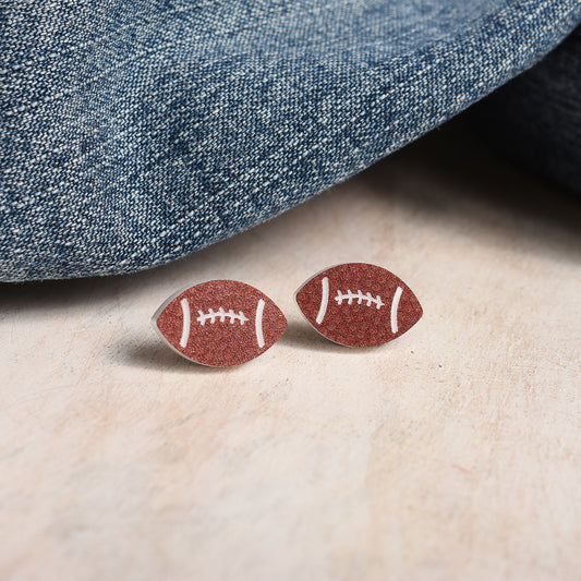 A pair of football-shaped stud earrings with a brown finish and white laces, displayed on a light beige surface next to a folded blue denim fabric.