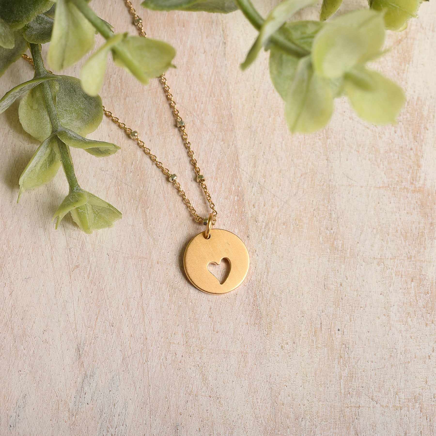 Gold necklace with a heart-shaped pendant on a light wooden surface with green leaves.