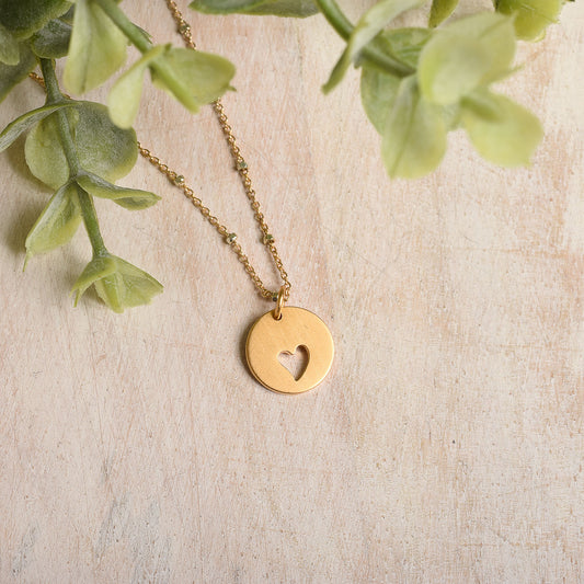 Gold necklace with a heart-shaped pendant on a light wooden surface with green leaves.