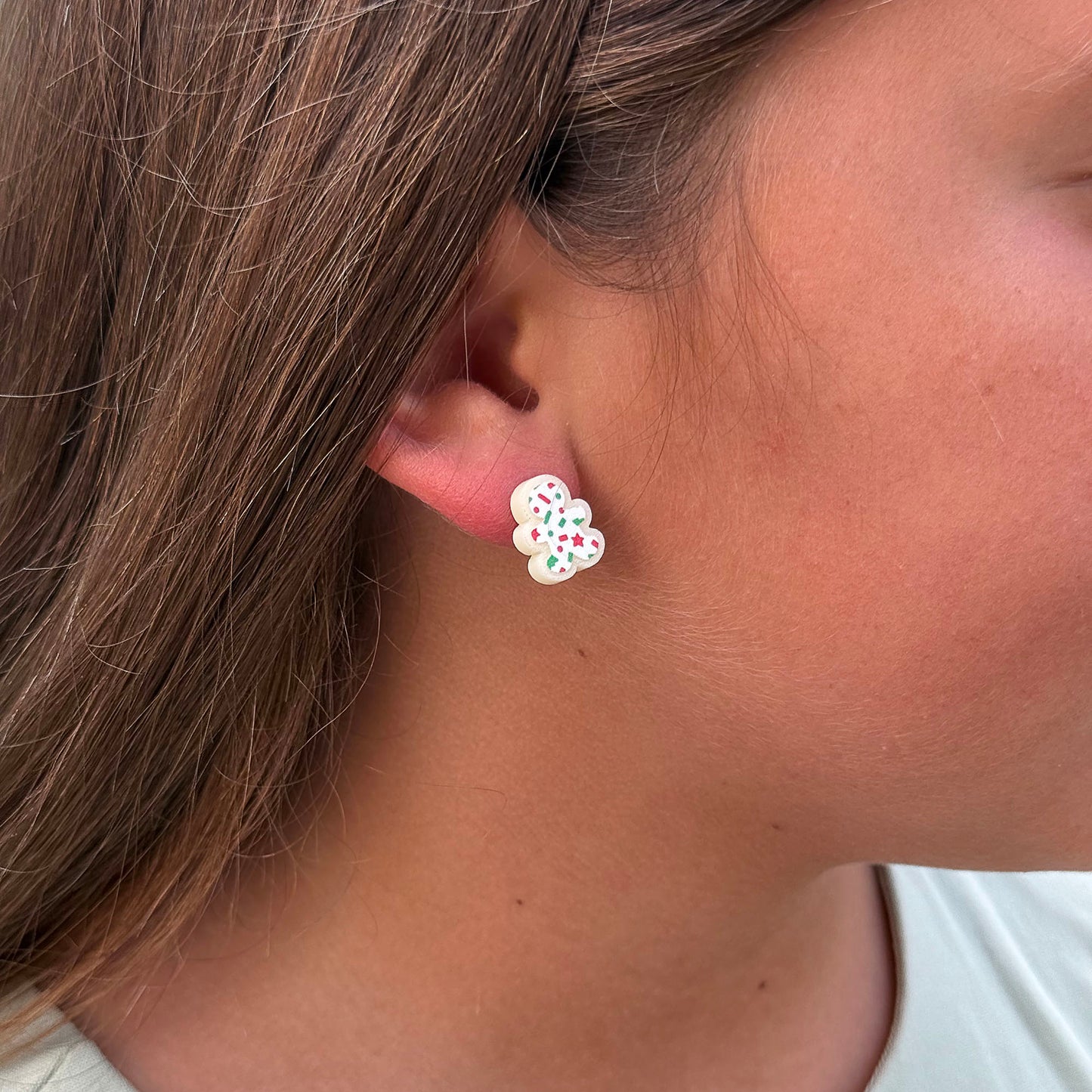 A close-up of a person's ear wearing a small gingerbread stud earring with green and red details.
