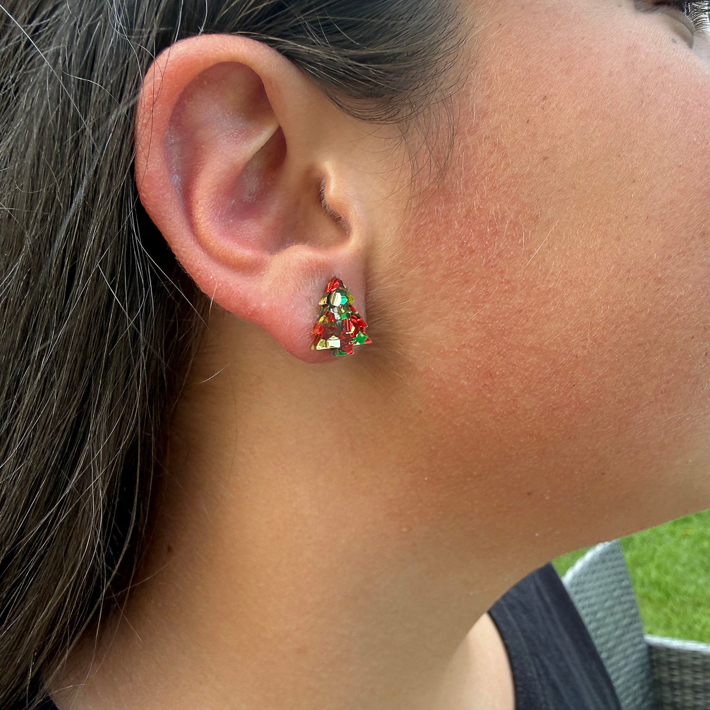 A close-up of a person's ear wearing a small Christmas tree stud earring with red, green, and gold details.