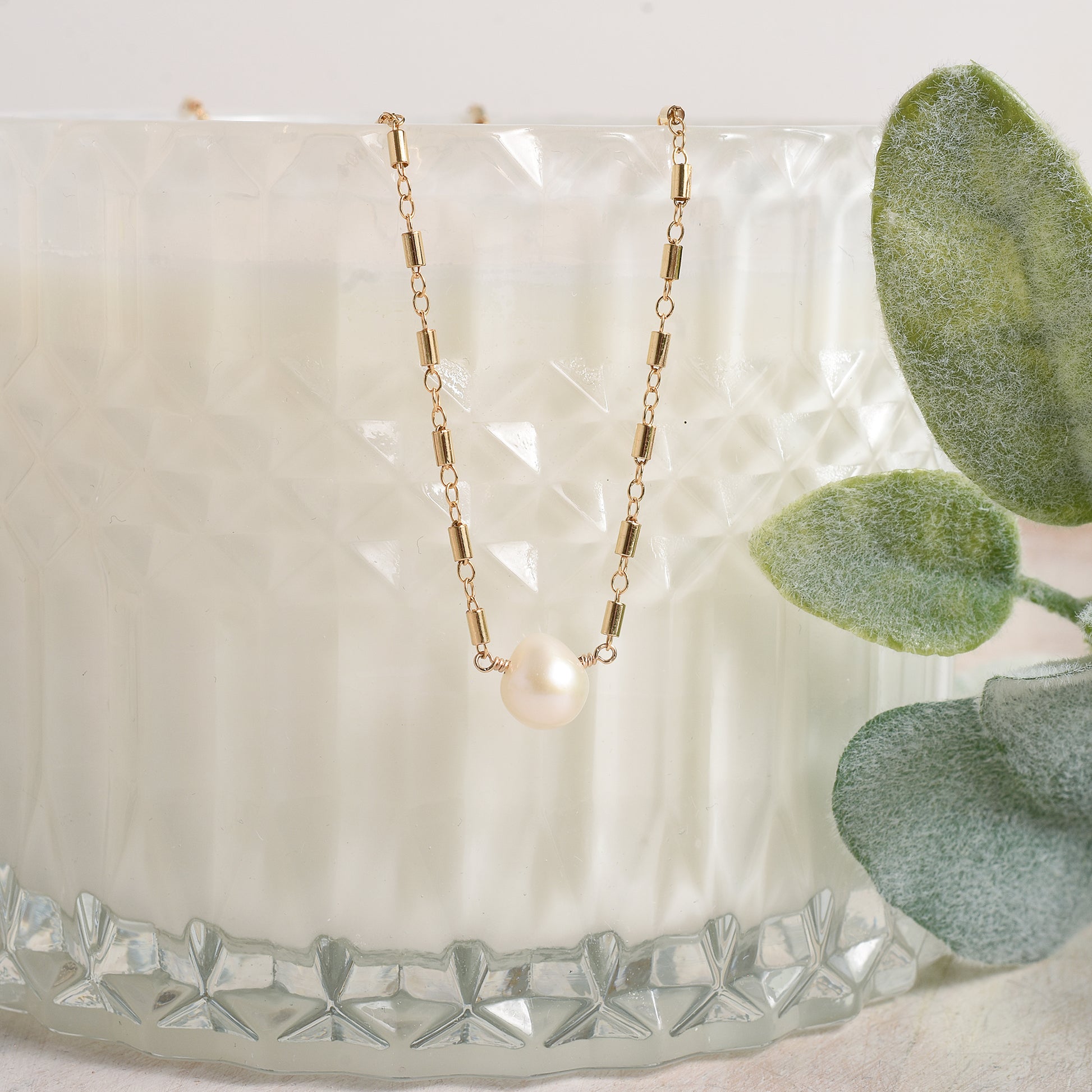 Gold necklace with a pearl pendant on a white surface with green leaves.