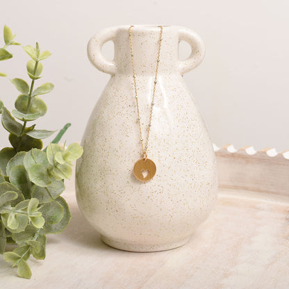 Necklace with a gold pendant hanging from a white textured vase on a light surface with greenery.