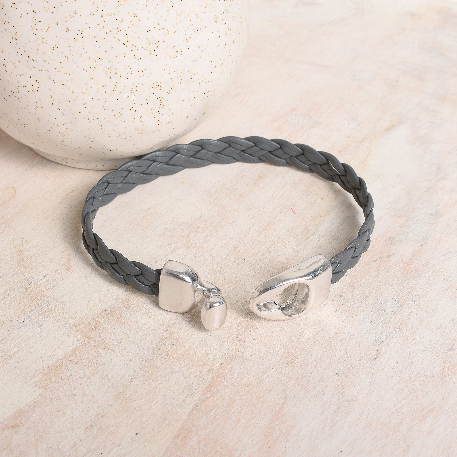 A gray braided leather bracelet with polished silver-tone toggle clasp, displayed on a light beige surface.