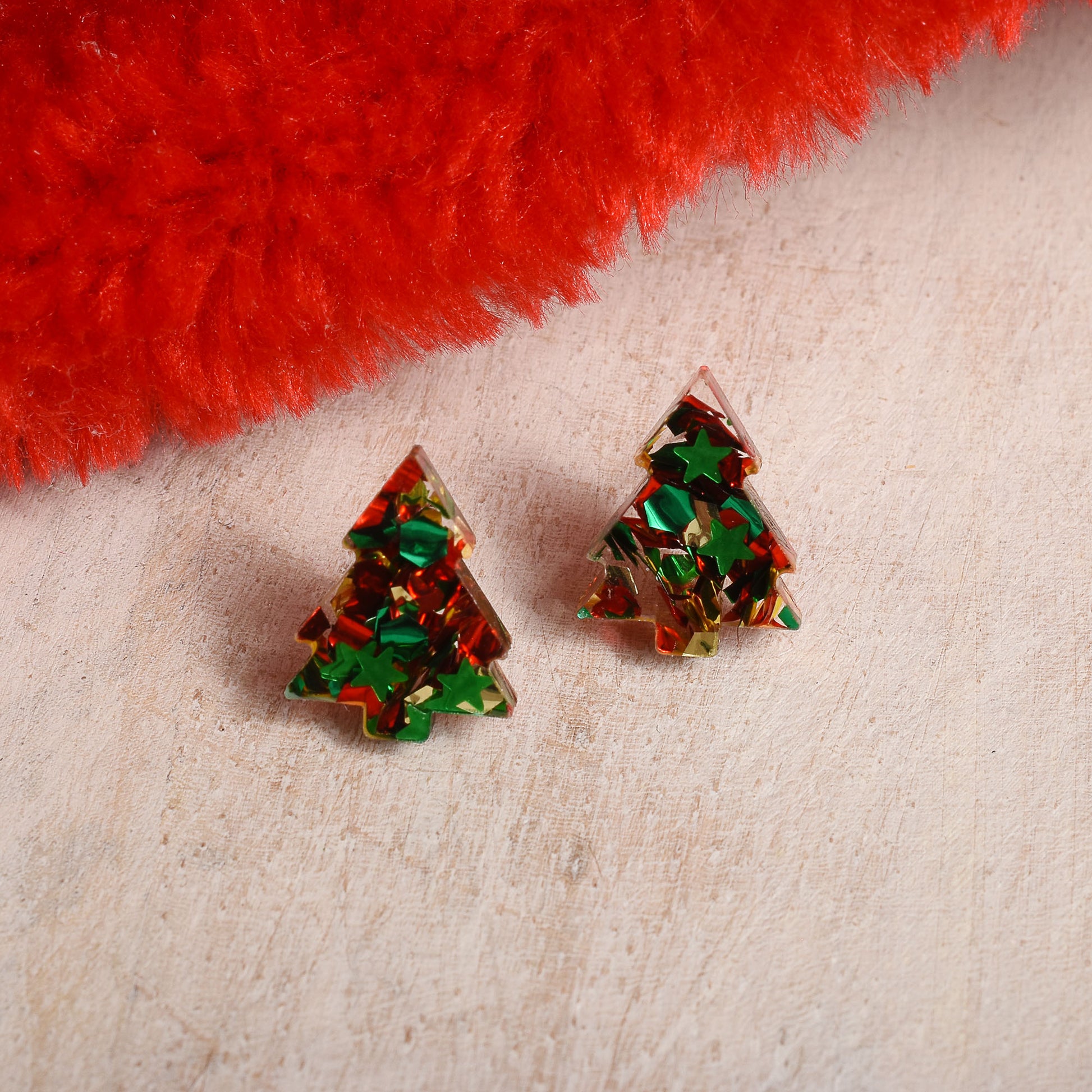 A pair of small tree-shaped stud earrings filled with iridescent multicolor glitter, resting on a textured beige surface with a bright red fuzzy prop at the top.