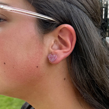 A close-up of a person's ear wearing a small glittery pink heart-shaped stud earring.
