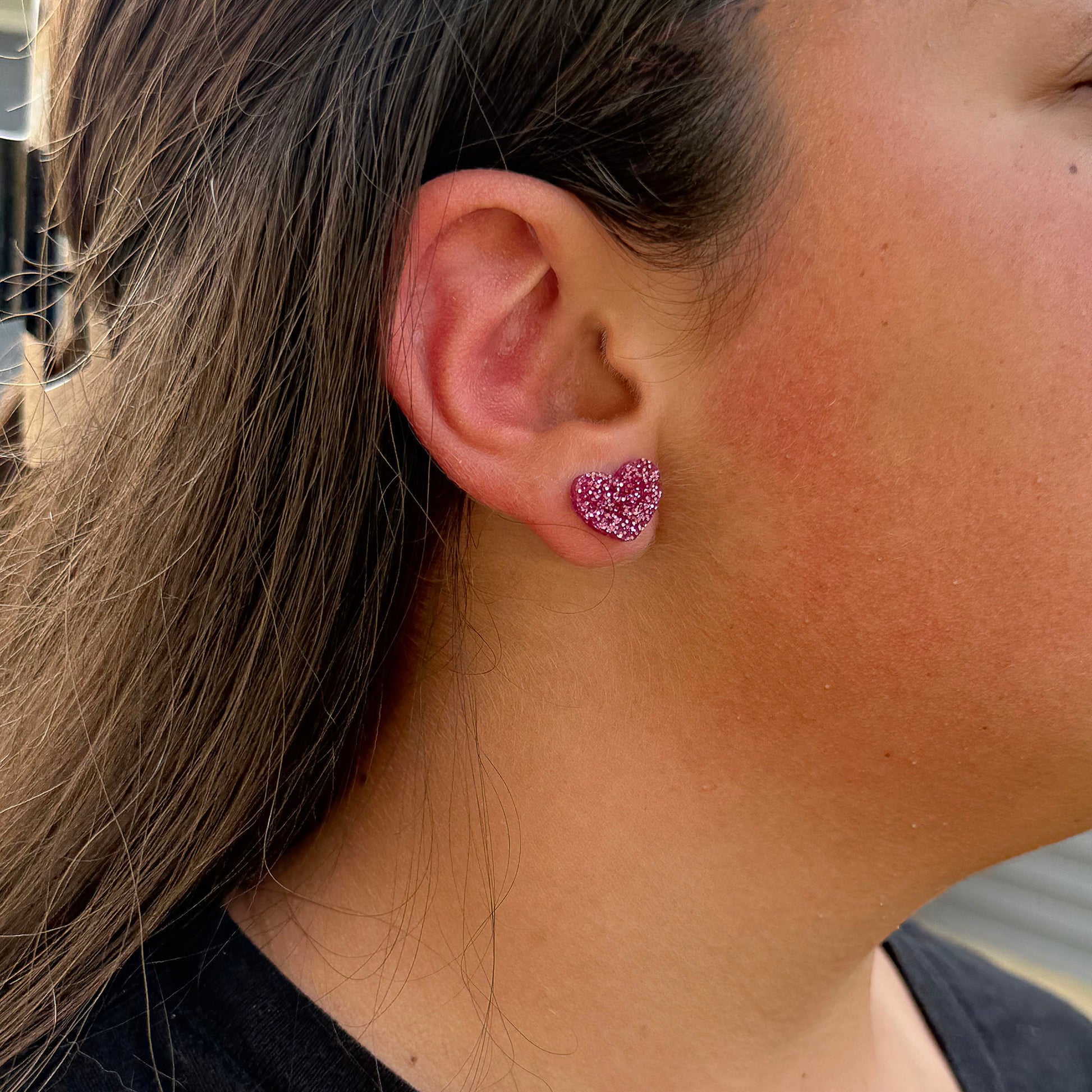 A close-up of a person's ear wearing a small glittery magenta pink heart-shaped stud earring.