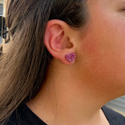 A close-up of a person's ear wearing a small glittery magenta pink heart-shaped stud earring.
