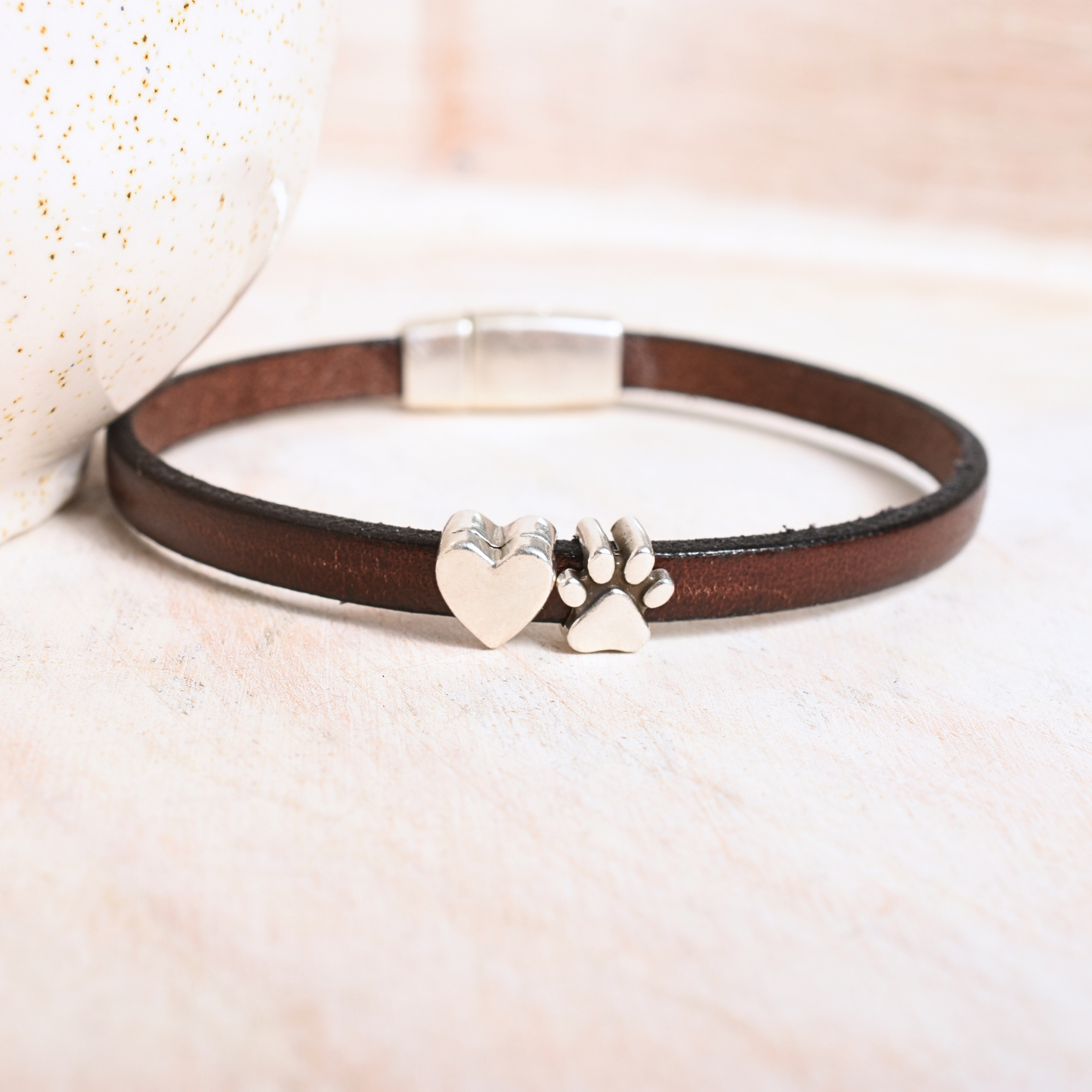Brown leather bracelet with silver heart and paw print charm on a soft beige background