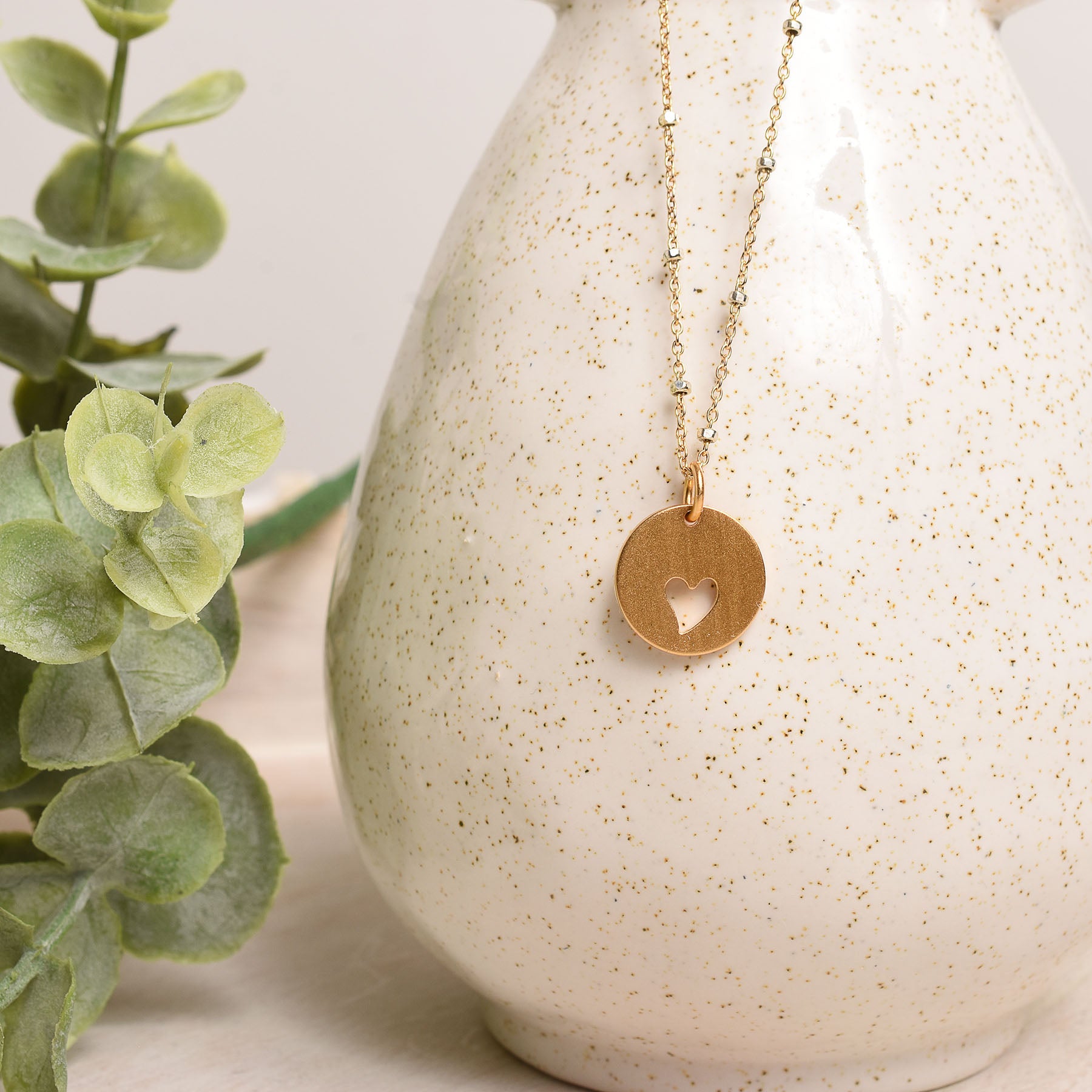 Gold necklace with a heart pendant on a textured white vase next to a green plant