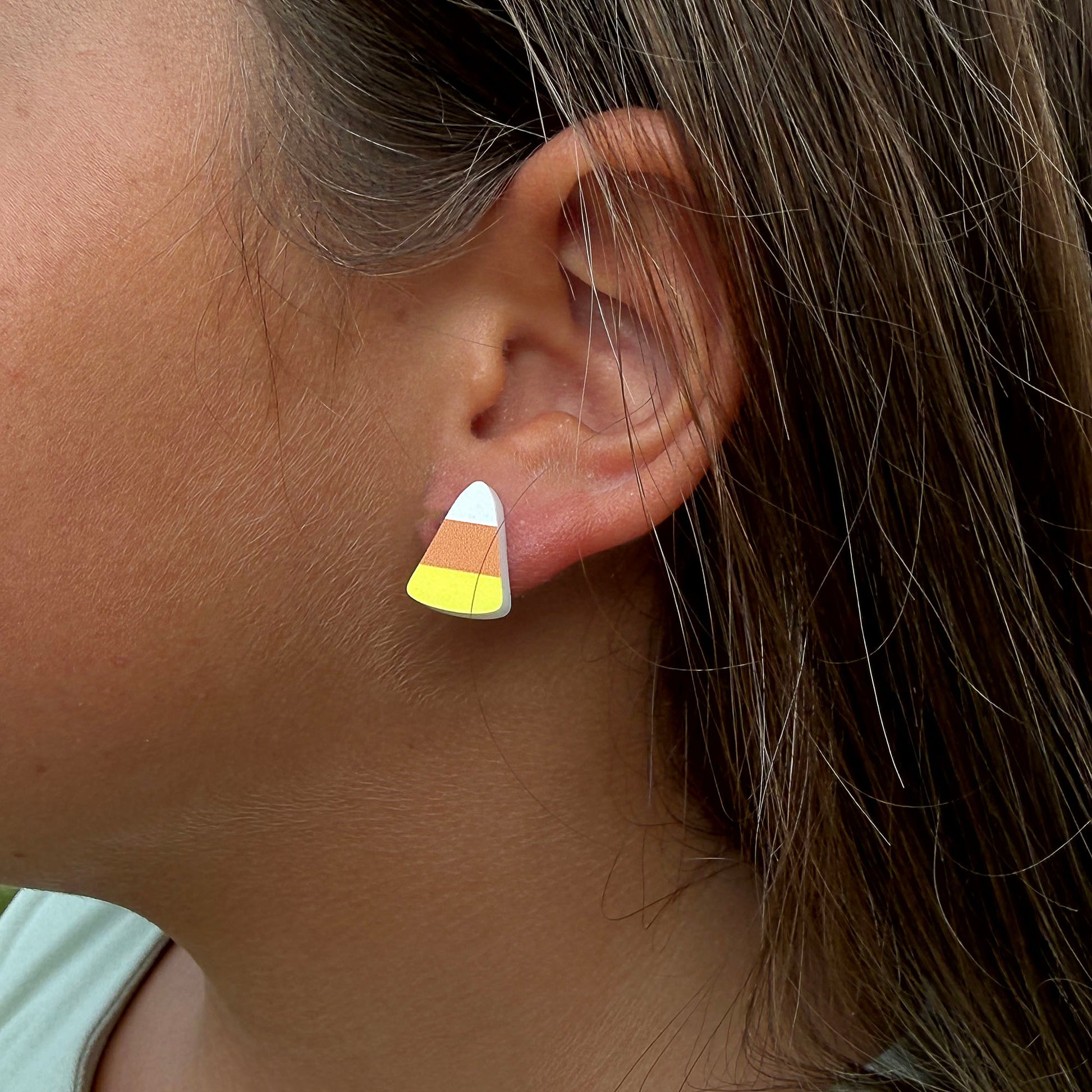 A close-up of a person's ear wearing a small stud earring shaped like a candy corn, with white, orange, and yellow sections.