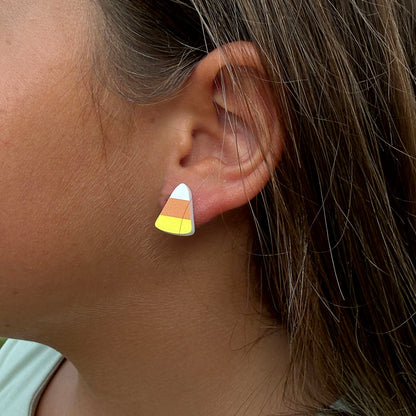 A close-up of a person's ear wearing a small stud earring shaped like a candy corn, with white, orange, and yellow sections.